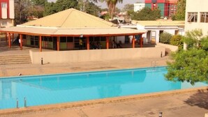 A swimming pool with a covered pavilion, a building with a red and white facade, and a clear sky. - Room-agura Hotel - Guest Room (Abuja)