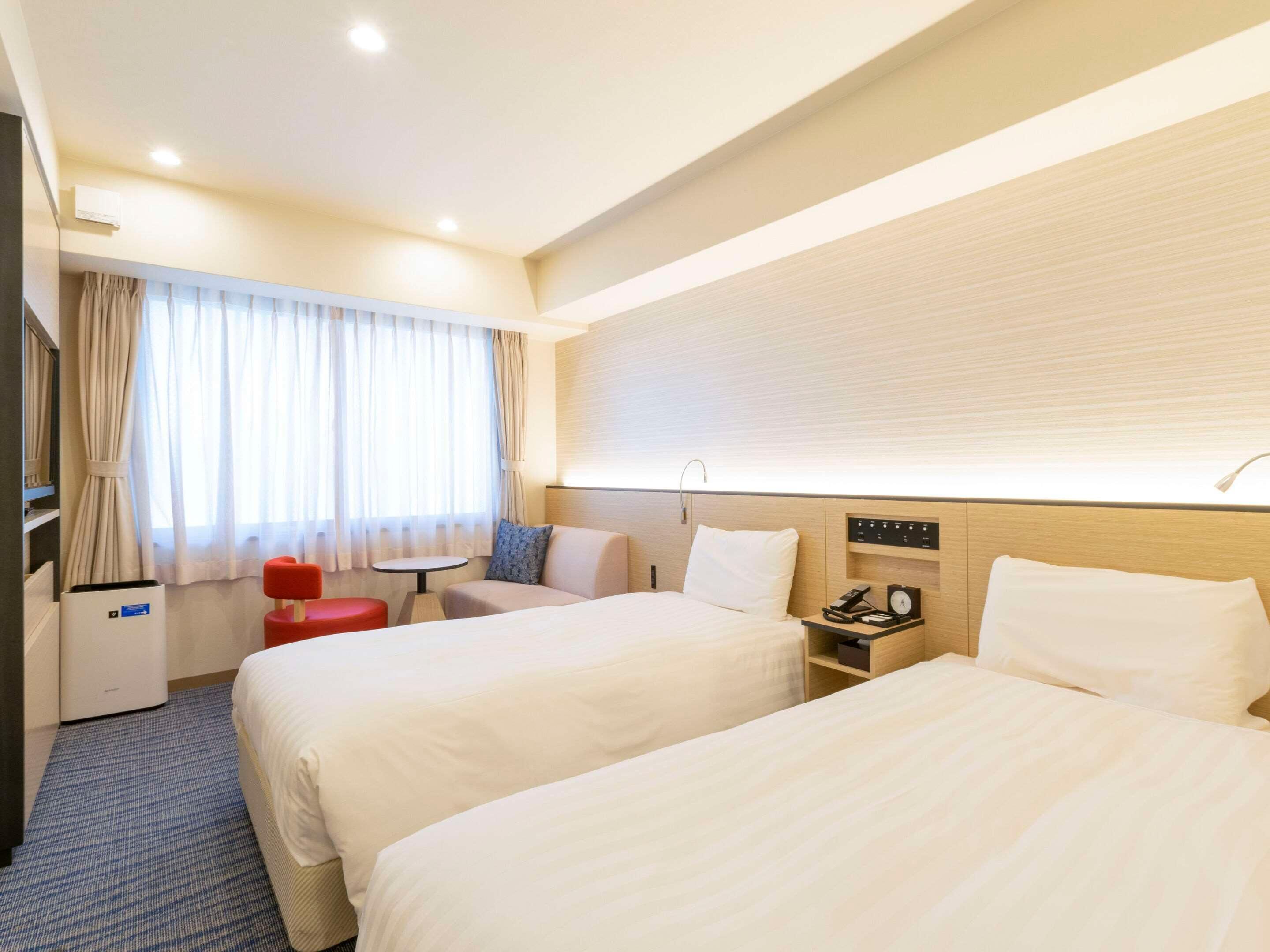 Une chambre d’hôtel avec deux lits, un bureau, une chaise, une petite table, un téléphone et une fenêtre avec des rideaux.