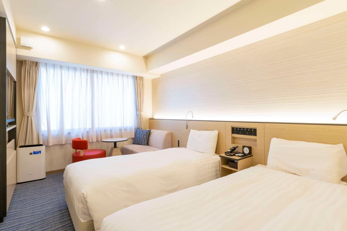A hotel room with two beds, a desk, a chair, a small table, and a window with curtains.