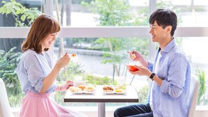 Dos personas sentadas a una mesa con comida, en una habitación con ventanas amplias y follaje visible desde el exterior.