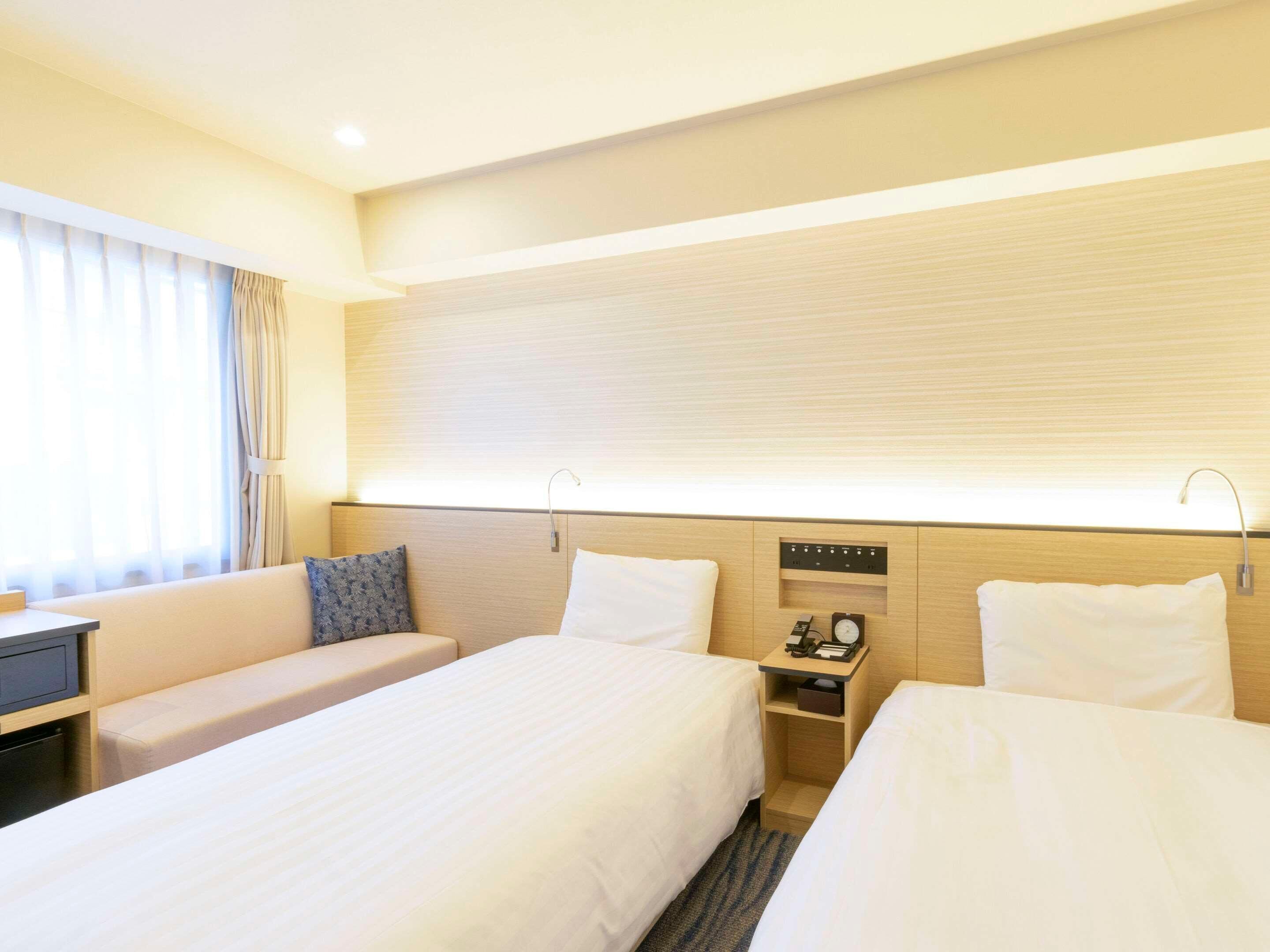 Une chambre d’hôtel avec deux lits, un bureau, une table de chevet avec une horloge et une fenêtre avec des rideaux.