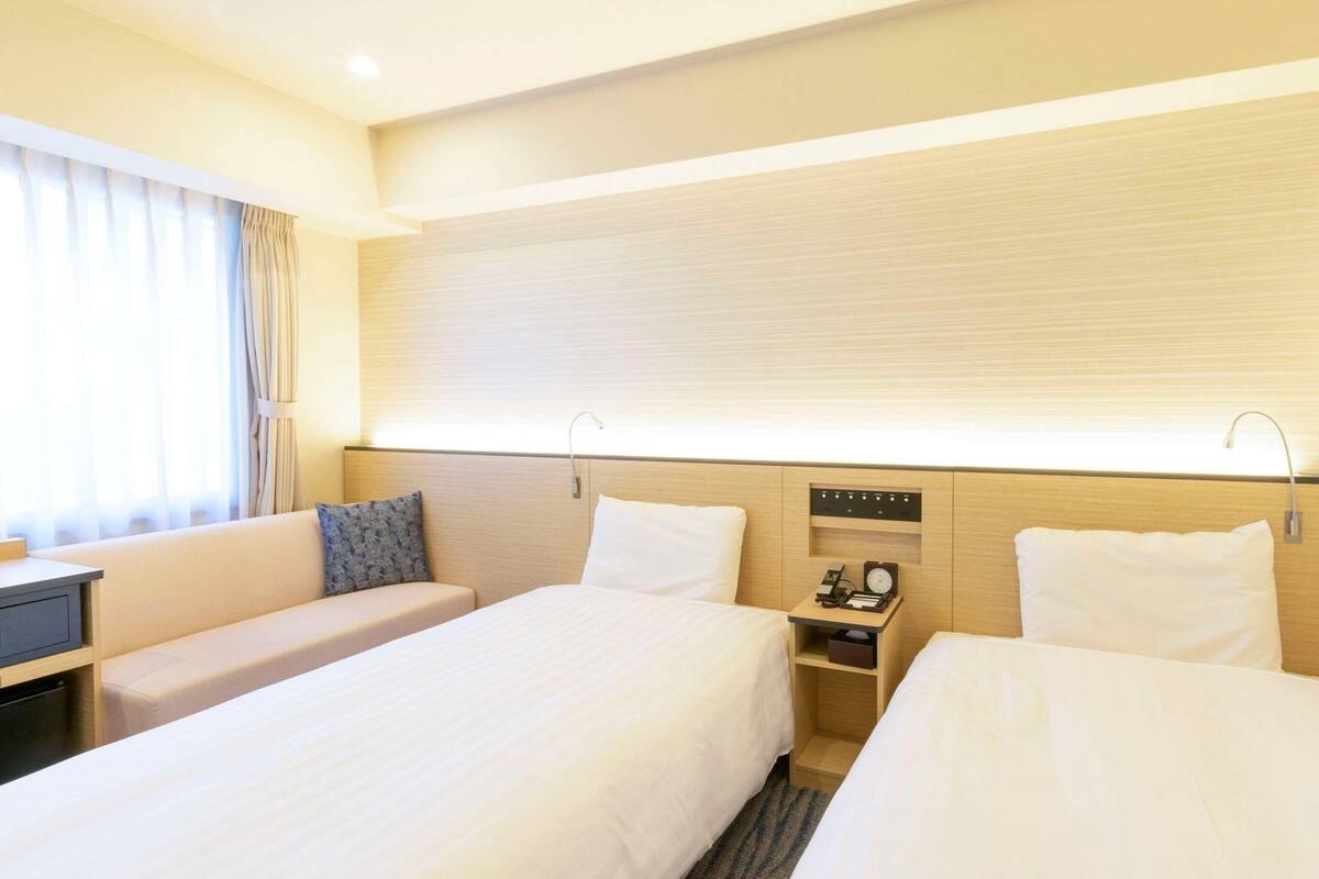 A hotel room with two beds, a desk, a nightstand with a clock, and a window with curtains.