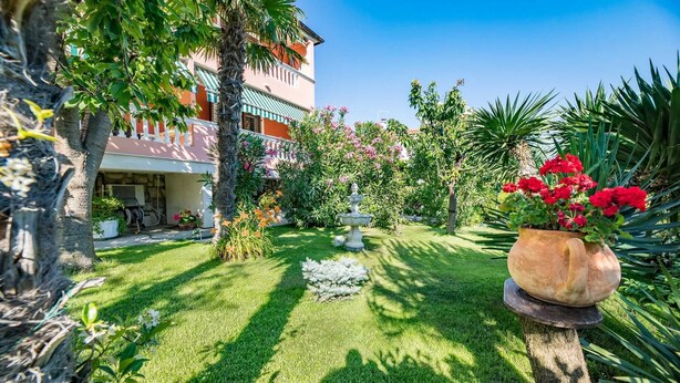 Um jardim bem cuidado com diversas plantas e flores, uma fonte de pedra e um edifício com varanda.