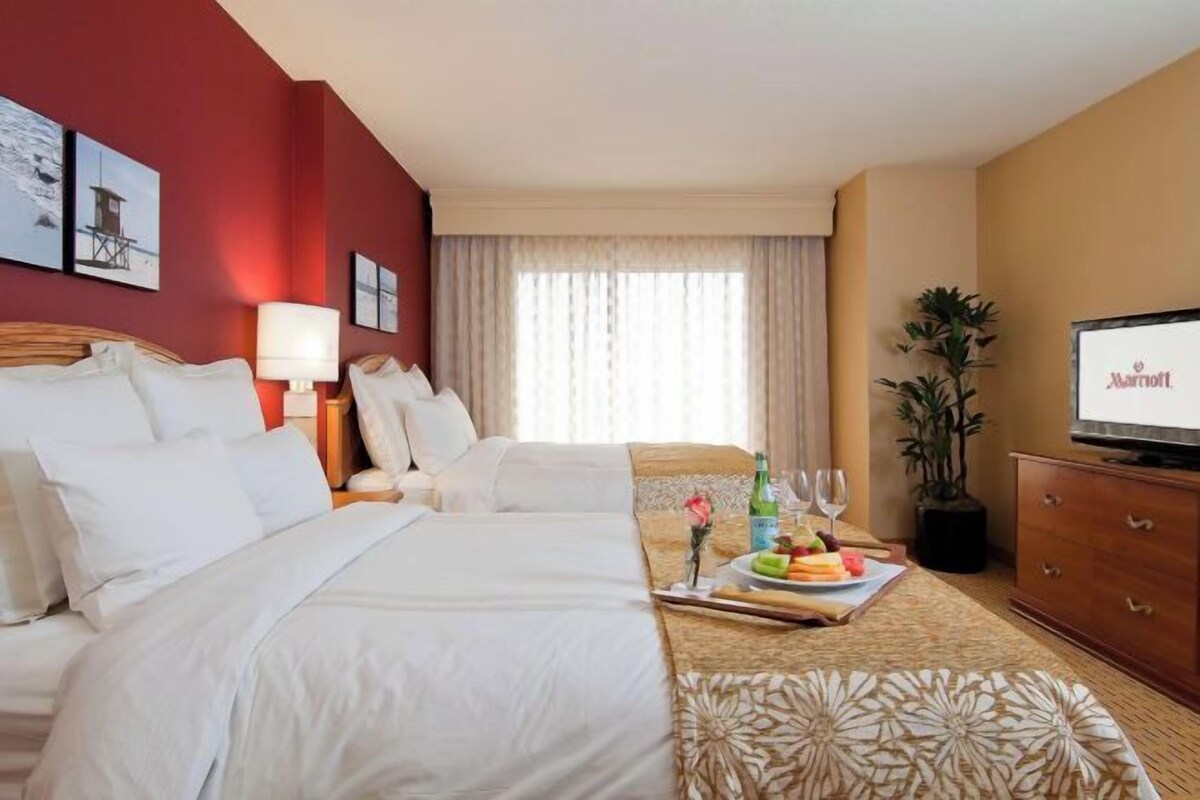 A hotel room with two beds, a television, a desk with a fruit plate, and a potted plant.