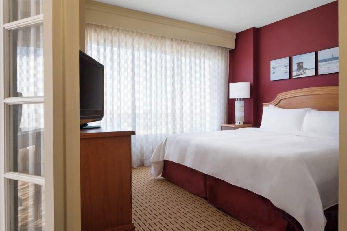 A hotel room with a bed, a television on a stand, a nightstand with a lamp, and a window with curtains.