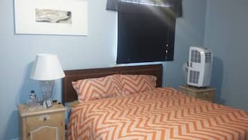 A bedroom with a bed, a nightstand, a lamp, a picture on the wall, and a standing fan.