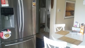 Een roestvrijstalen koelkast met waterdispenser en vriesvak. Er staat een klein tafeltje met een geweven tafelmat en twee witte stoelen. Een schilderij hangt aan de muur, en een deur is open naar een andere kamer.