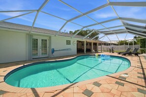 Une piscine avec un patio couvert, un bâtiment blanc doté d’une porte et une cour clôturée.