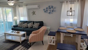 Ein Wohnzimmer mit einem dunkelgrünen Sofa, einem kleinen, pfirsichfarbenen Sessel, einem weißen Couchtisch, einem Esstisch mit gewebten Untersetzern und einem Deckenventilator.