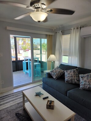 Un salon doté d’un ventilateur de plafond, d’un canapé agrémenté de coussins à motifs, d’une table basse et d’une porte-fenêtre coulissante donnant sur un espace extérieur avec une table et des chaises.