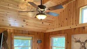 Interior acogedor de una cabaña con paredes y techo de madera, dos sofás con diversos cojines, un ventilador de techo y un ventanal con vista a árboles.