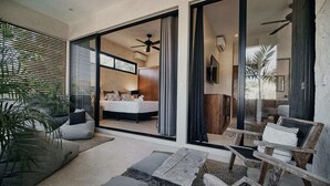 Ein modernes Wohnzimmer mit einer Couch, einem Couchtisch und einem Holzstuhl. Eine große Schiebeglasdoor führt in ein Schlafzimmer mit einem Bett und einem Deckenventilator.