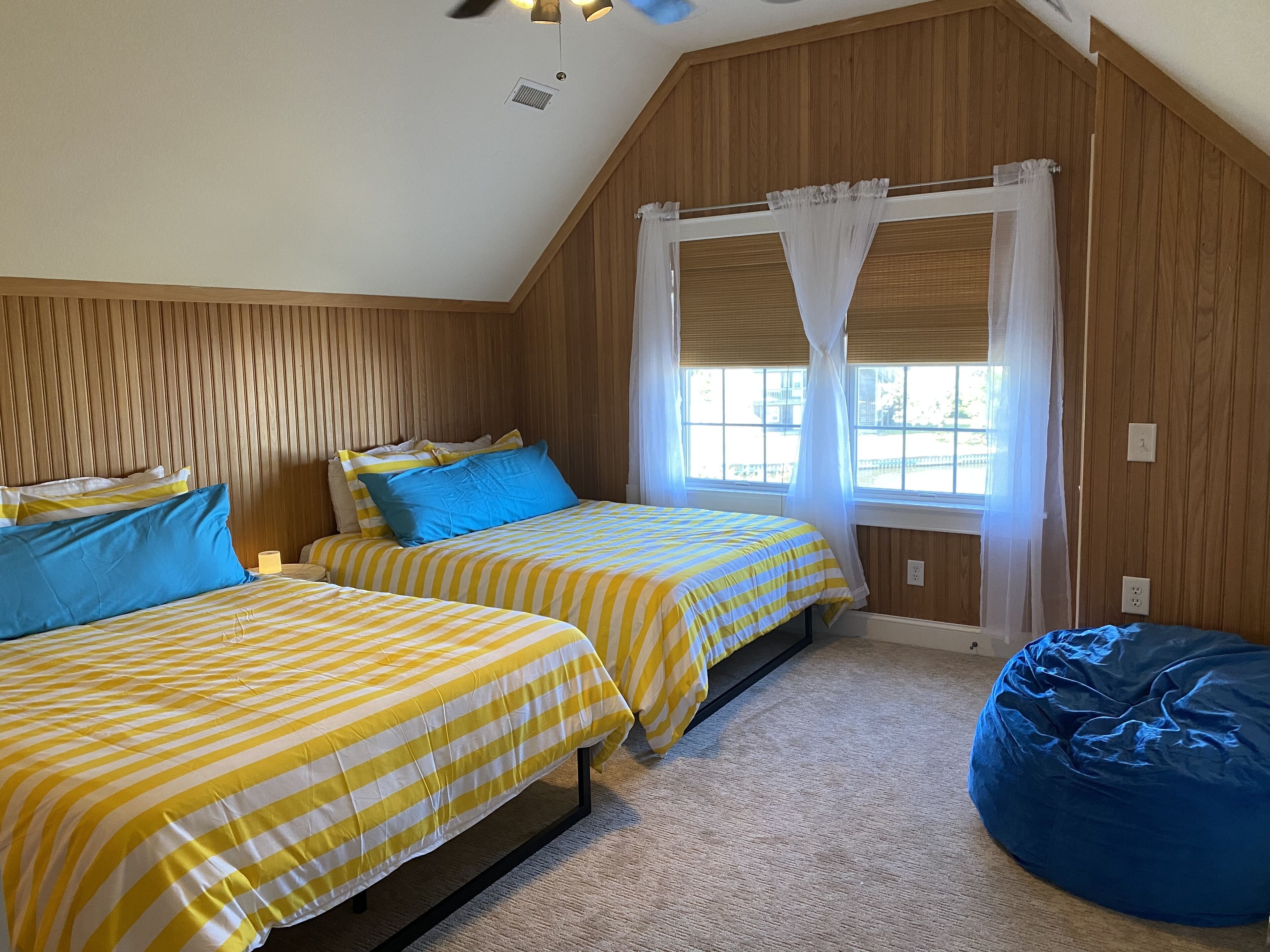 A bedroom with two beds, a window with curtains, a ceiling fan, and a bean bag chair.