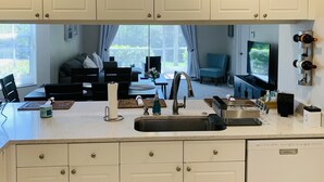 A kitchen with white cabinets, a built-in sink, a ceiling fan, and a view to a living area with a TV and seating.