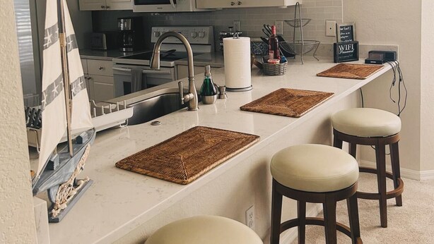 A modern kitchen with white cabinets, a microwave, and a sink. There are three stools and a small sailboat decoration.