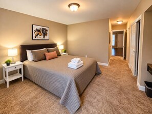 Quarto de hotel com uma cama grande, duas mesas de cabeceira, um criado-mudo, uma obra de arte na parede e piso coberto por carpete.