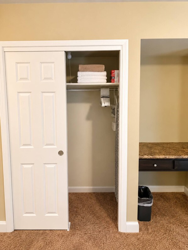 A room with a closed door, a closet with folded towels, a shelf with a box, and a trash bin.