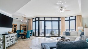 A spacious living room with a large flat-screen TV, a ceiling fan, and a dining area with blue chairs and a wooden cabinet.
