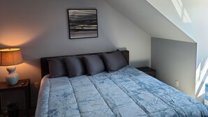 Et soveværelse med skråvægge, en seng med et blåt tæppe, et natbord med en lampe og et billede i ramme på væggen.