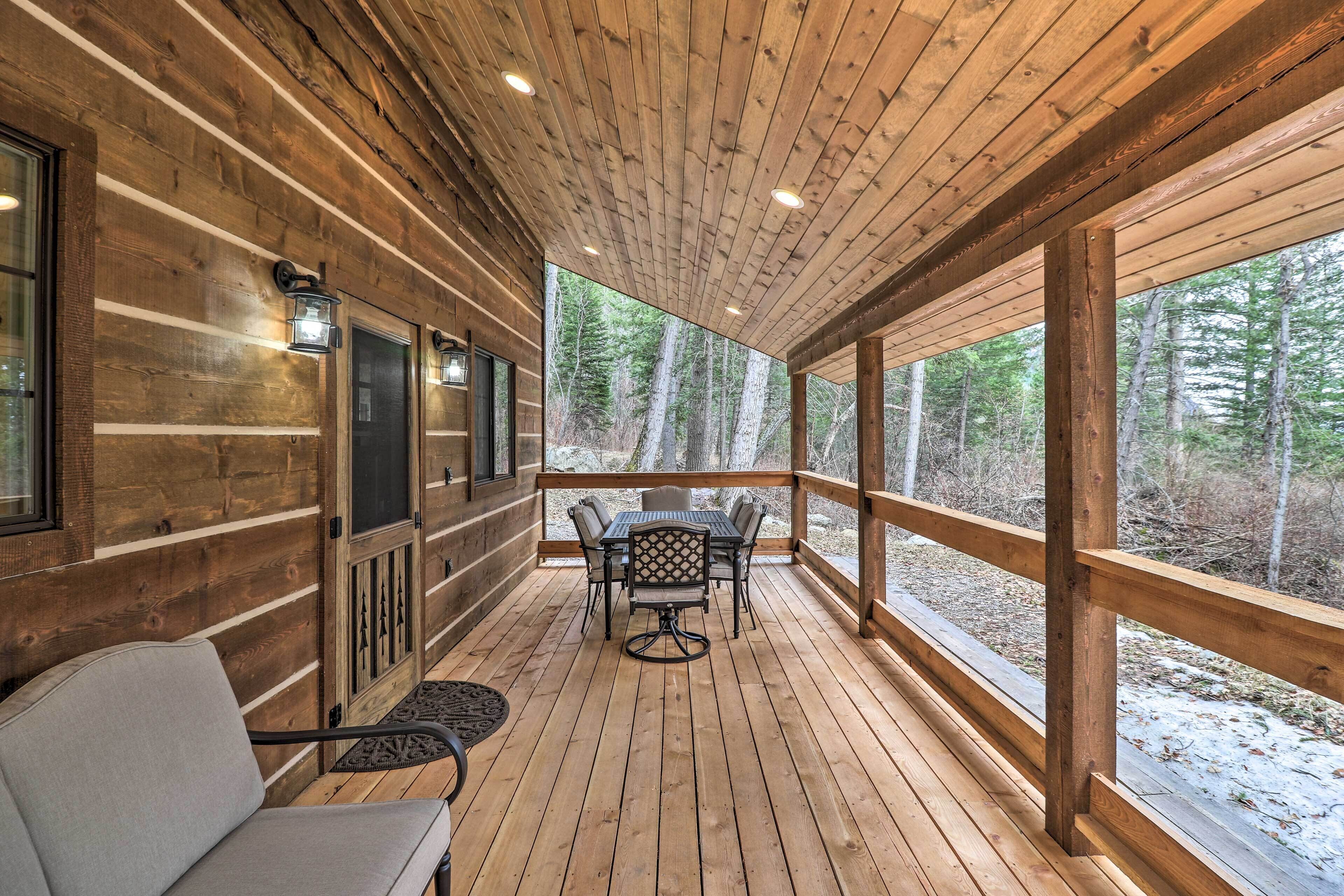 A wooden deck with a table and chairs, surrounded by large windows offering a view of a forested area.