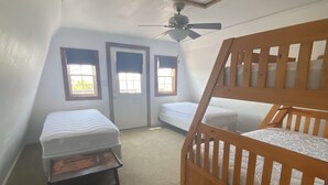 Une petite chambre mansardée de forme triangulaire, avec deux lits simples, un ventilateur de plafond et deux fenêtres équipées de rideaux.