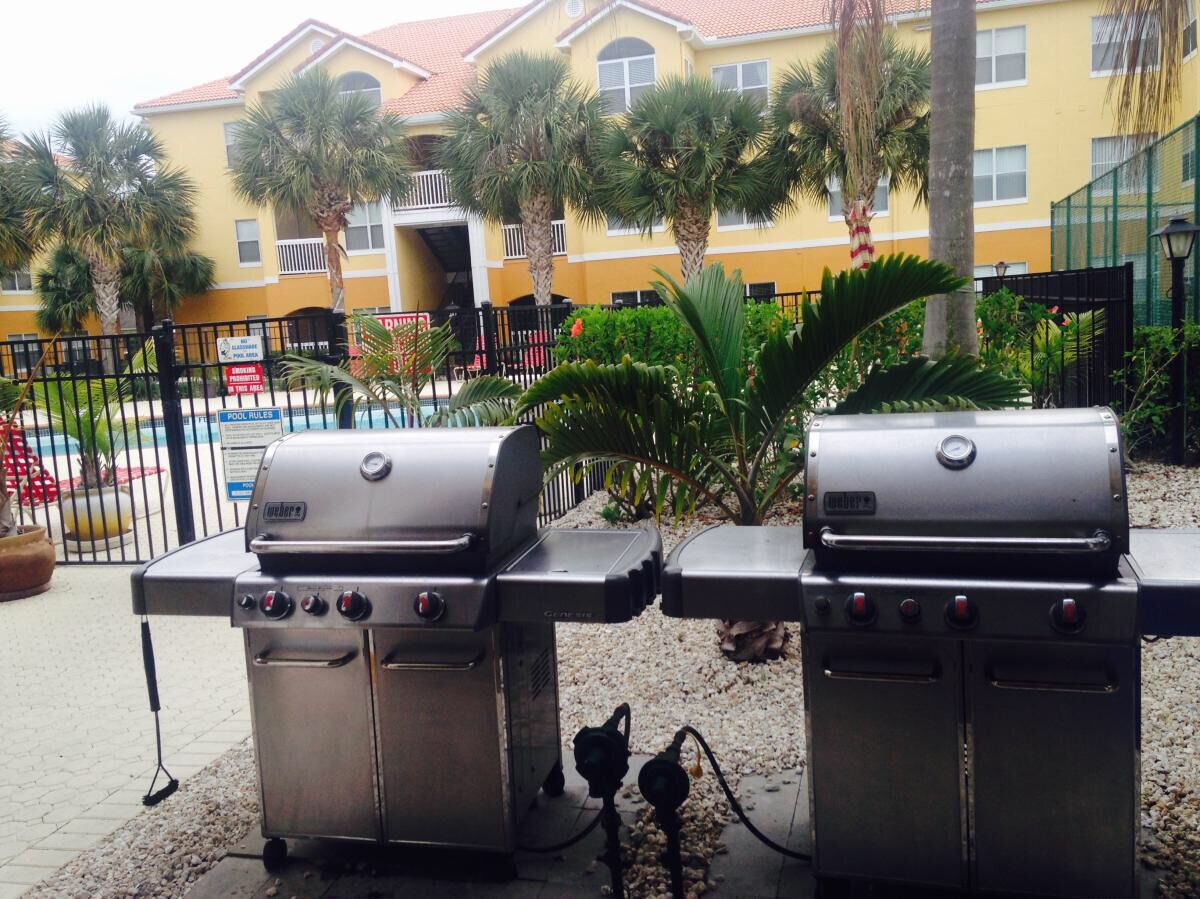Two outdoor grills in a courtyard with a pool and a yellow building in the background.