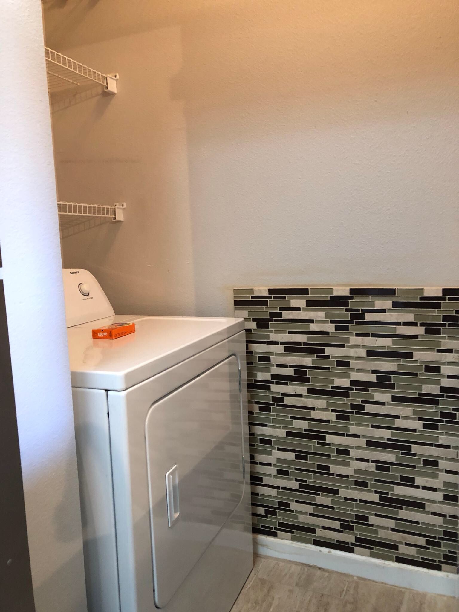 A laundry area with a front-loading washer and dryer, a tiled backsplash, and a white wall.