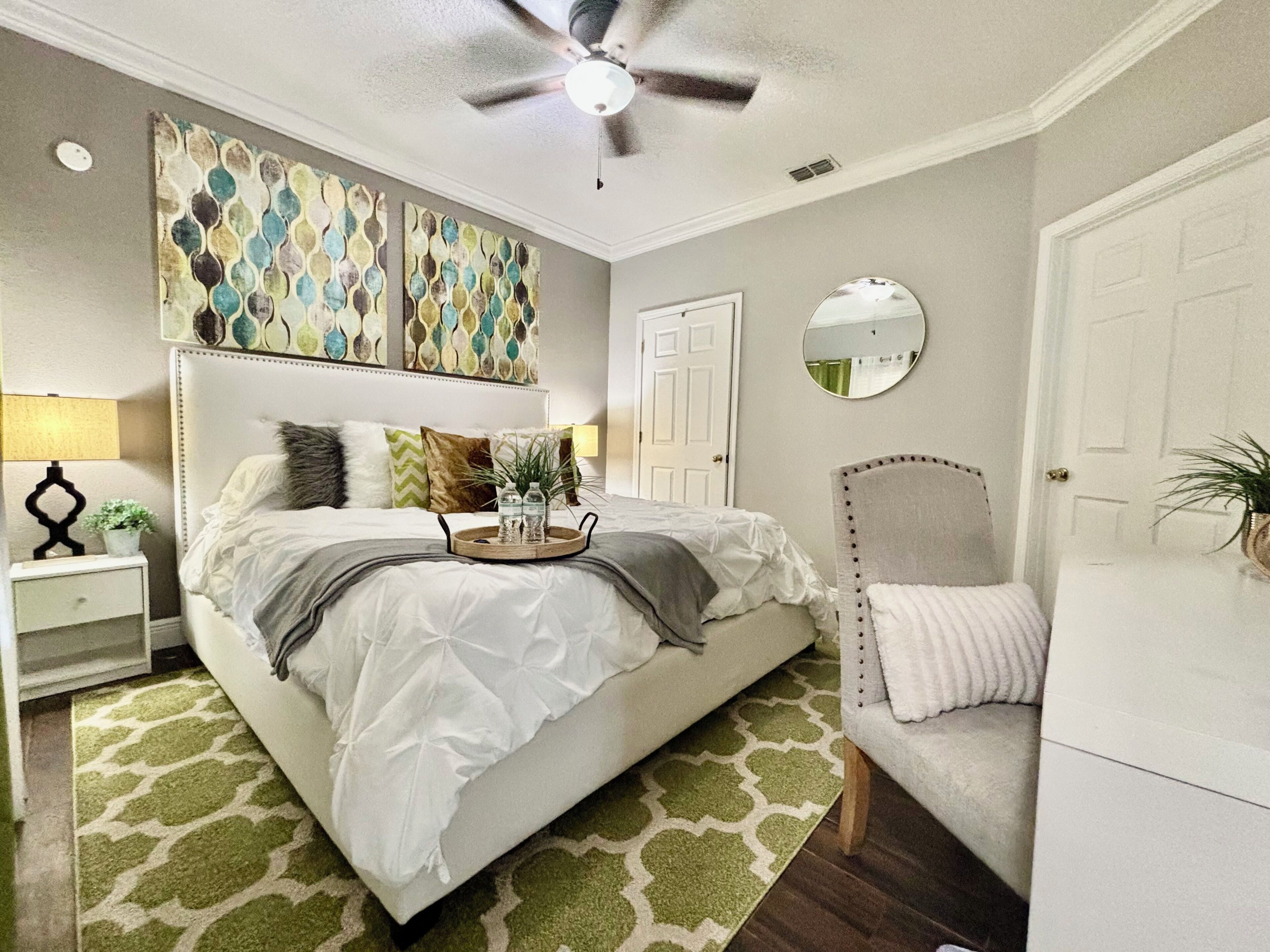 Un dormitorio con cama, mesita de noche, lámpara, almohadillas decorativas, obra de arte, ventilador de techo, espejo y puerta.