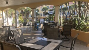 Een terras buiten met tafels en stoelen, omgeven door palmbomen en een gebouw op de achtergrond.