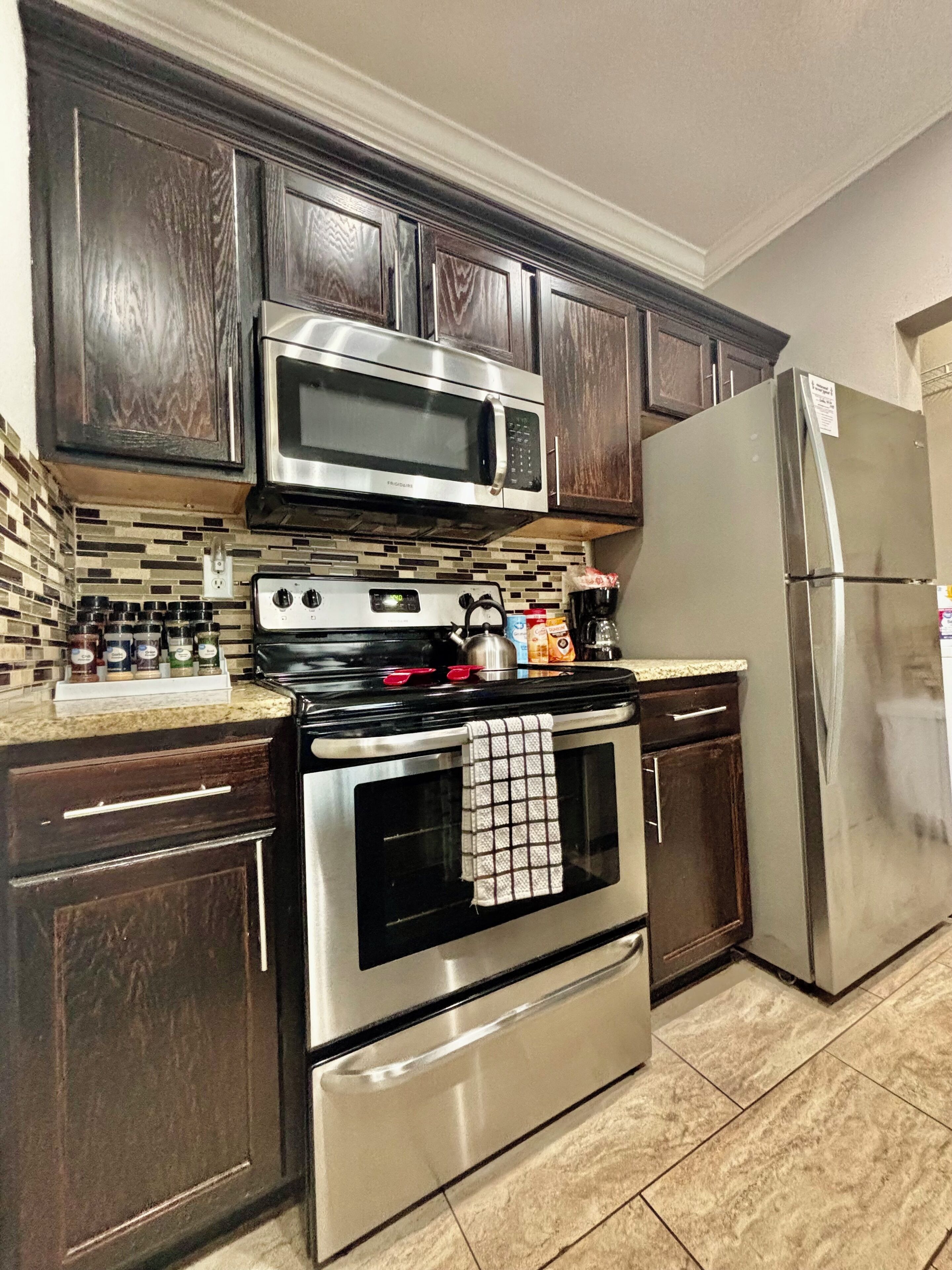 Una cocina con armarios de madera oscura, electrodomésticos de acero inoxidable, incluyendo un microondas, estufa y refrigerador, y salpicadero de azulejos.