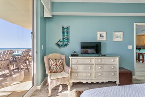 Una habitación con un ventanal grande que da a una terraza, una silla de mimbre con cojín, un aparador blanco y un televisor.