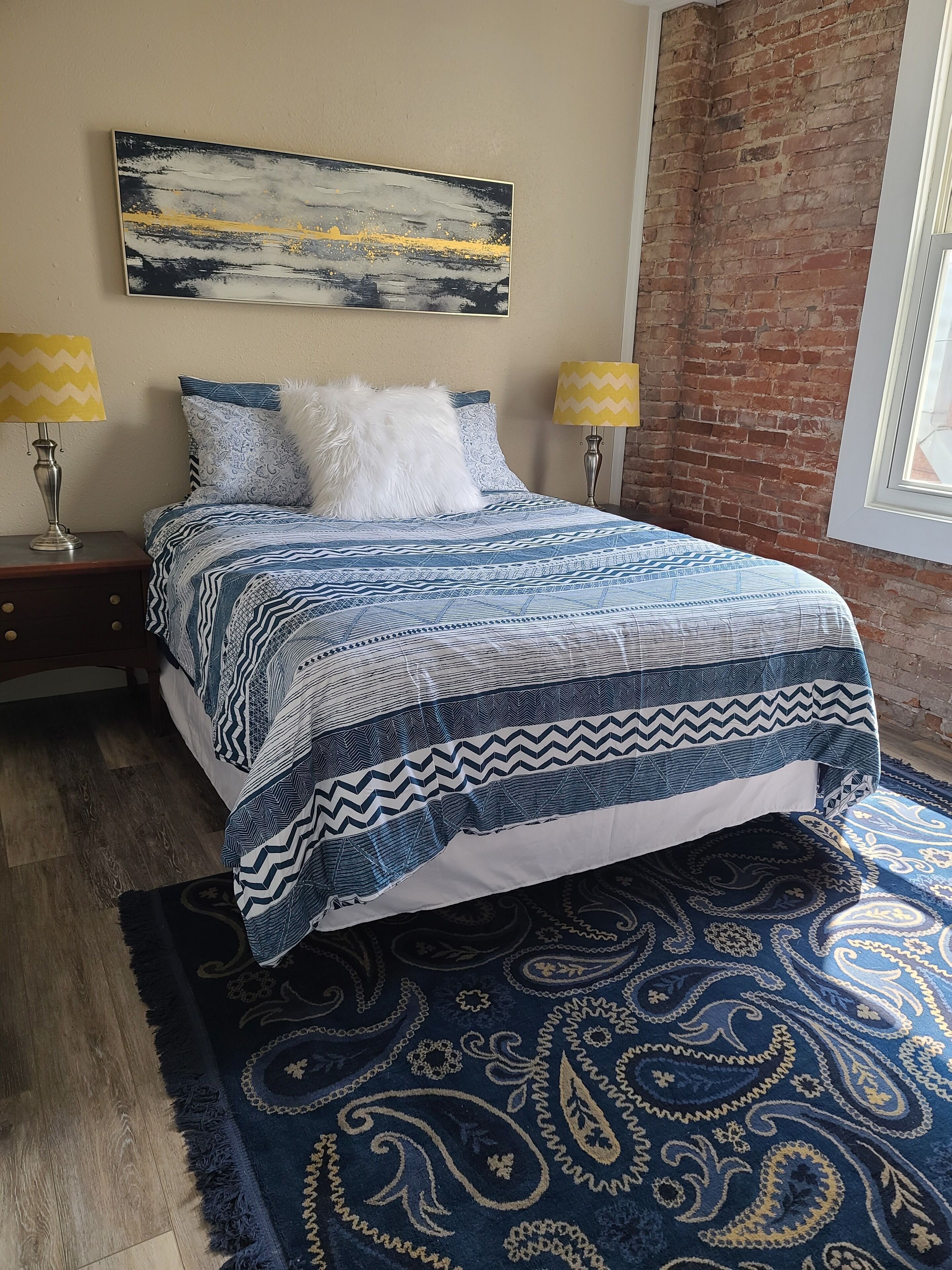 A bed with a patterned bedspread, two bedside lamps, a wall-mounted abstract painting, and a patterned rug.