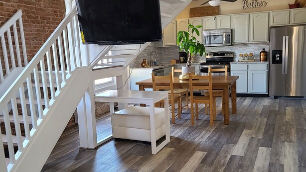 A spacious living area with a wooden floor, a dining table with chairs, a flat-screen TV mounted on the wall, and a kitchen area with modern appliances. - The Lofts of North Main Street (Saint Charles)