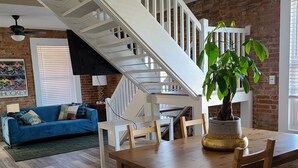 A spacious living area with a wooden dining table, a blue sofa, and a staircase with white railings. - The Lofts of North Main Street (Saint Charles)