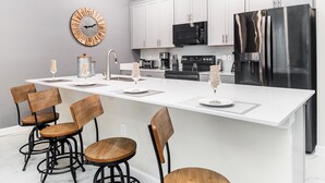 A modern kitchen with white cabinetry, a stainless steel refrigerator, a microwave, and a large island with bar stools.