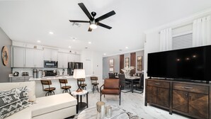 A modern living room with a sofa, coffee table, and TV, connected to a kitchen with white cabinets and a dining area with chairs.