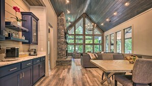 A spacious kitchen with dark cabinets, a stone fireplace, and a large dining area with a high ceiling and large windows. - Perfect for Couples,2 King Master bedrooms, Dry Sauna, Hot Tub, 3 covered decks (Broken Bow)