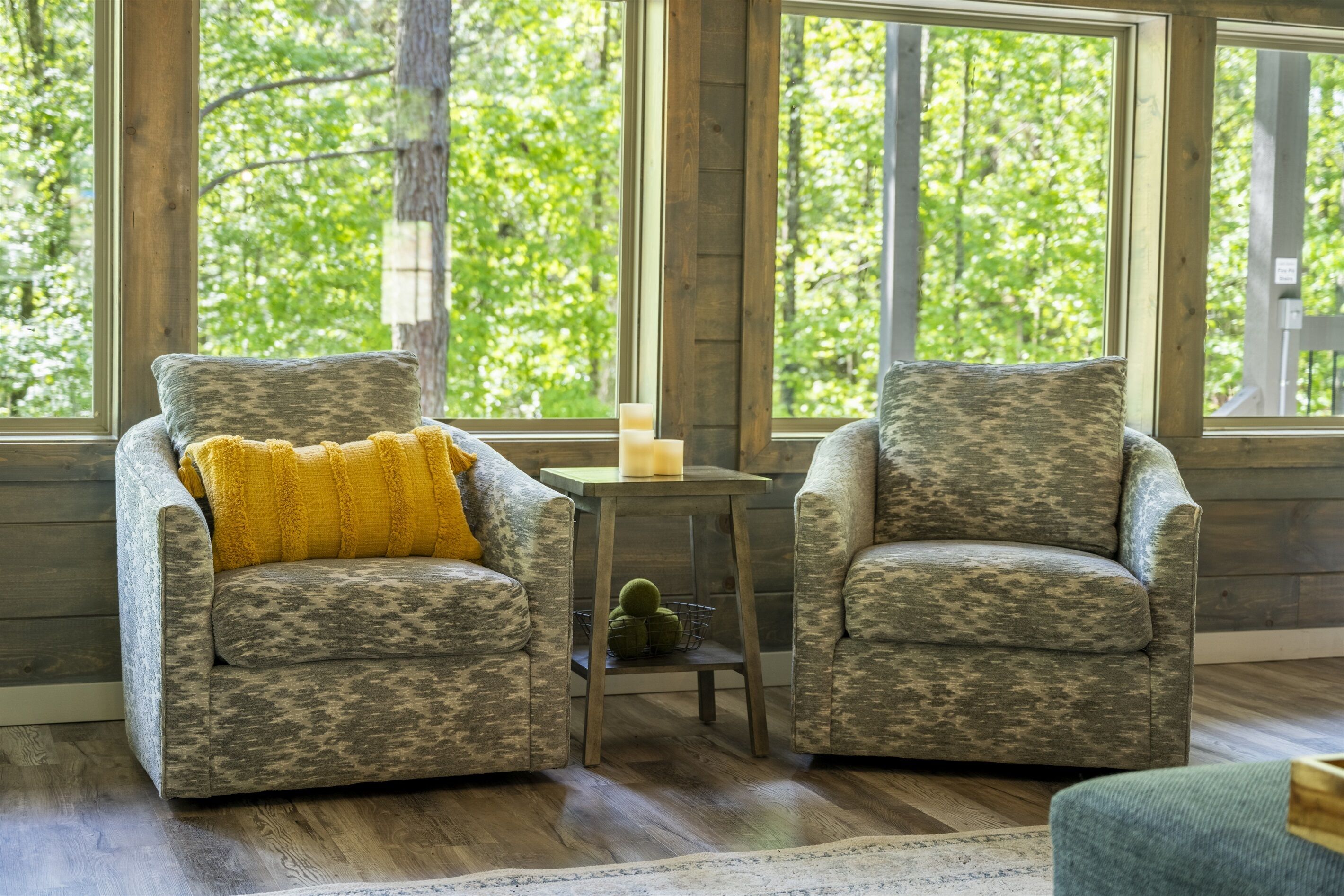 A room with two patterned armchairs, a small table with a candle, and a view of green trees through large windows.