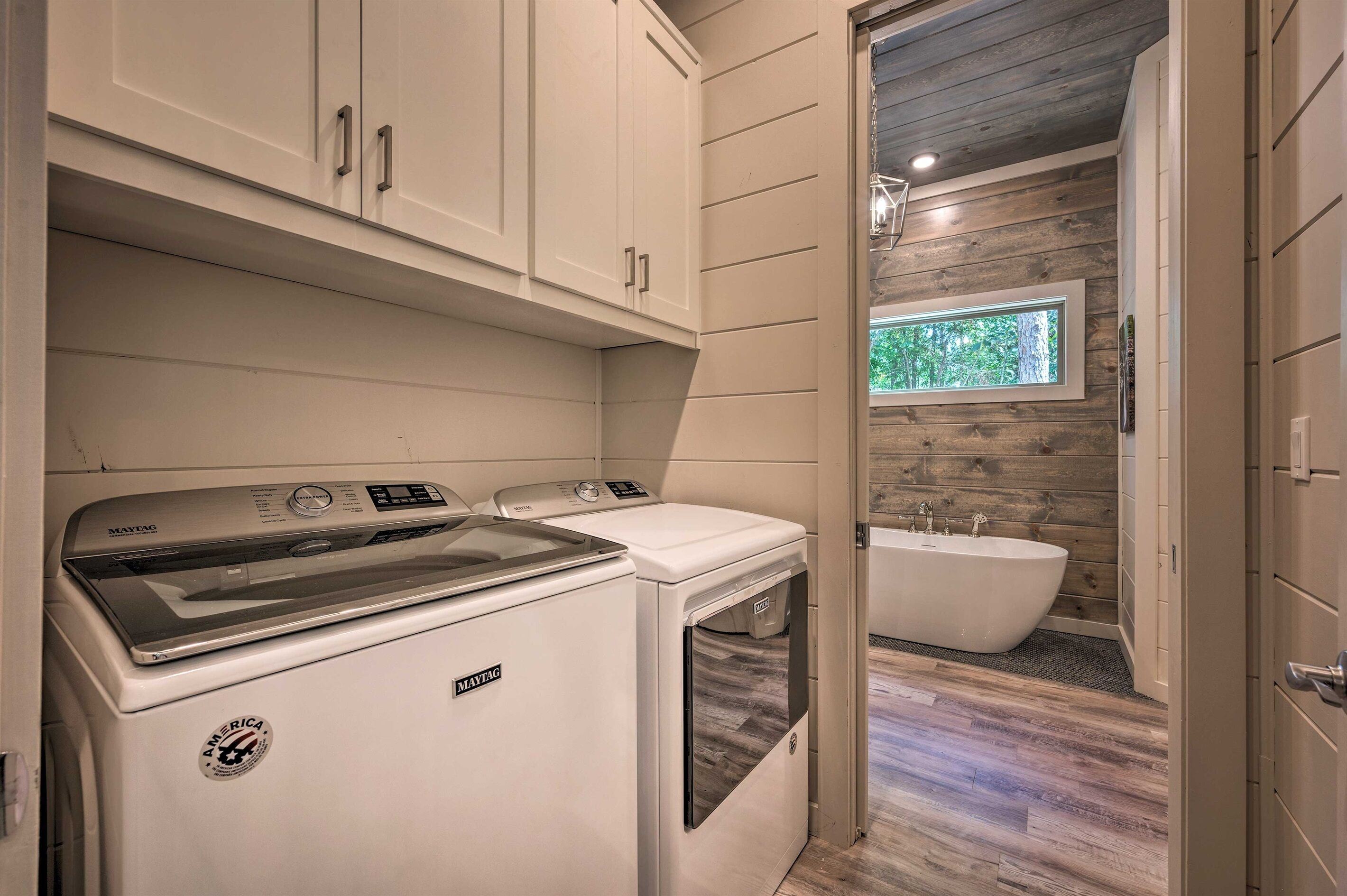A laundry room with a Maytag washer and dryer, a bathtub, and a window with a view of trees.
