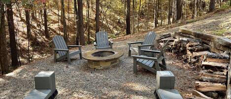 Un camping rodeado de bosque con un hoyo para la fogata, cuatro sillas Adirondack y un montón de leña.