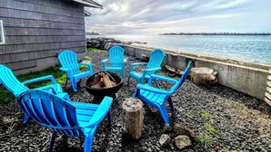 Ein Sitzbereich am Wasser mit blauen Adirondack-Stühlen, die um eine Feuerstelle angeordnet sind, mit Blick auf ein Gewässer.