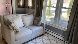 A light-colored sofa with two pillows, a patterned throw, and a fluffy rug in a room with a large window, a mirror, and a door. - Cute, Convenient and Clean (Wautoma)