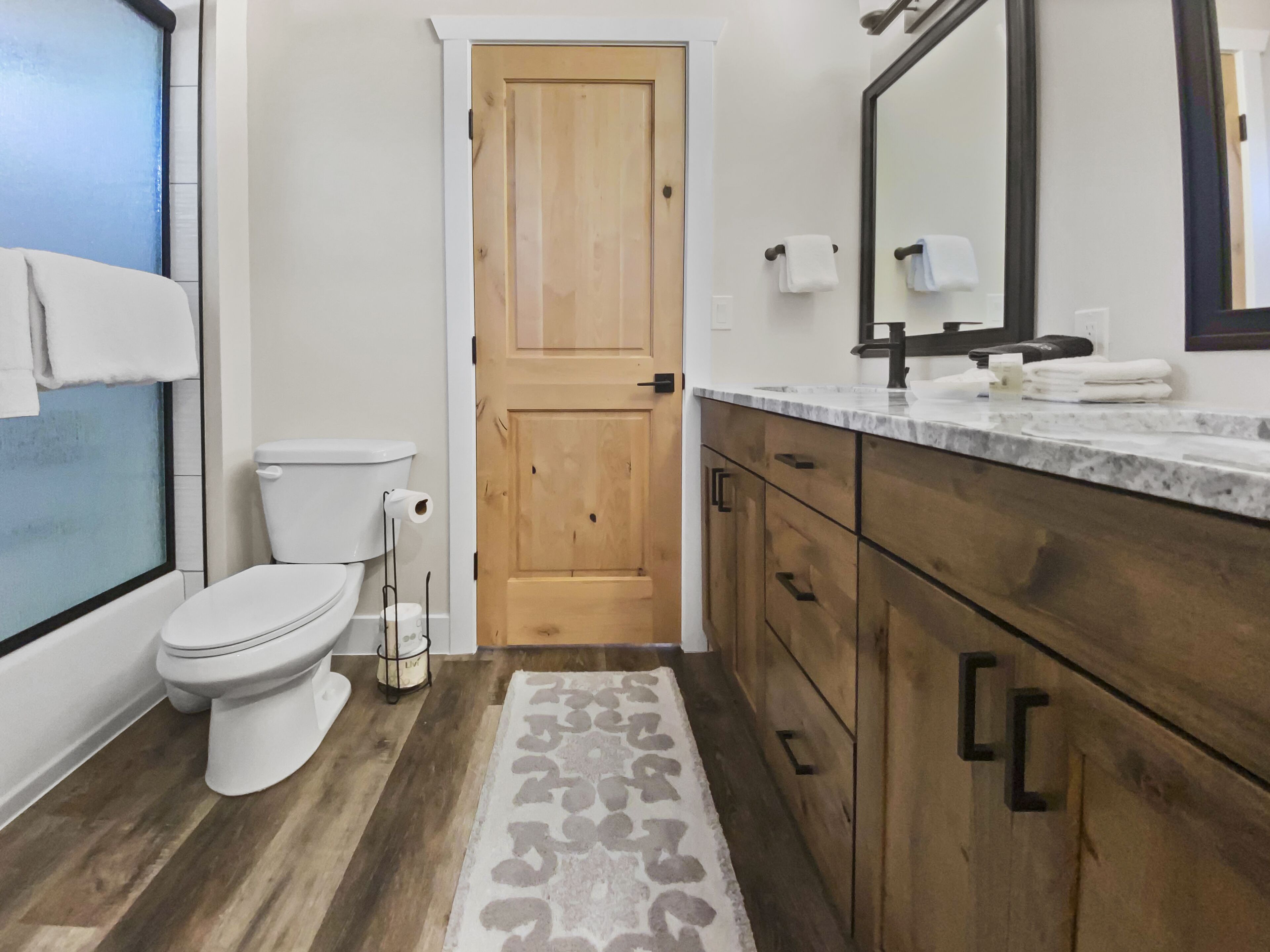 A bathroom with a toilet, wooden door, vanity with a mirror, and a patterned rug.