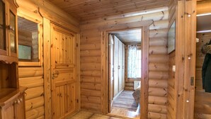 Interior de uma cabana de tronco com paredes de madeira, piso ladrilhado e corredor que leva a outro cômodo.