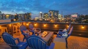 Un bar sul tetto con posti a sedere all'aperto, tra cui sedie blu e un braciere, con vista sullo skyline della città al crepuscolo.
