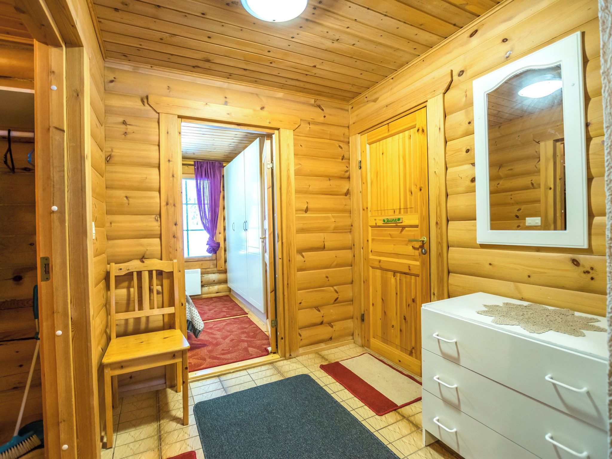 Interior de una cabaña de madera con una silla de madera, un aparador blanco y un espejo en la pared.