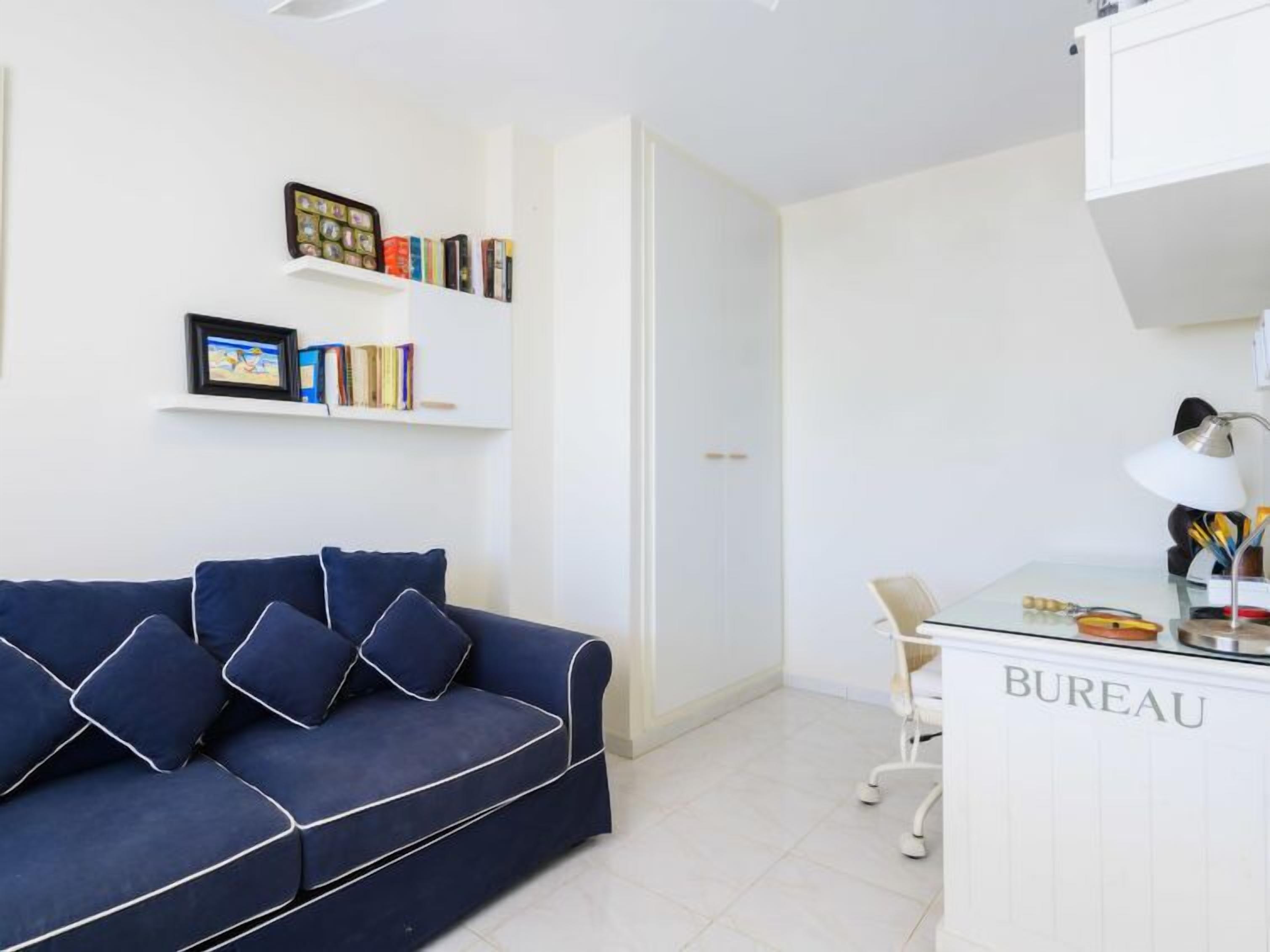 Una stanza moderna e minimalista con un divano blu scuro, una scrivania bianca con l'etichetta 'BUREAU' e mensole con libri e quadri incorniciati.