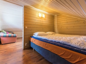 A small, cozy room with a wooden ceiling and walls, a single bed with a blue mattress, and a pink chair with a plaid cushion. - Rinteenkotka mökki by Interhome (Hyrynsalmi)