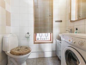 A compact bathroom with a toilet, a window with blinds, a washing machine, and a sink. - Rinteenkotka mökki by Interhome (Hyrynsalmi)