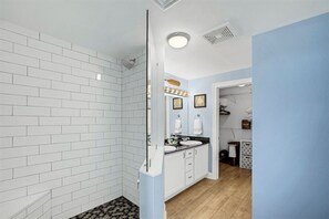 Ein Badezimmer mit weißen Fliesen, einer Dusche, einem Waschbecken mit Spiegel und einem Waschtisch mit Schränken.